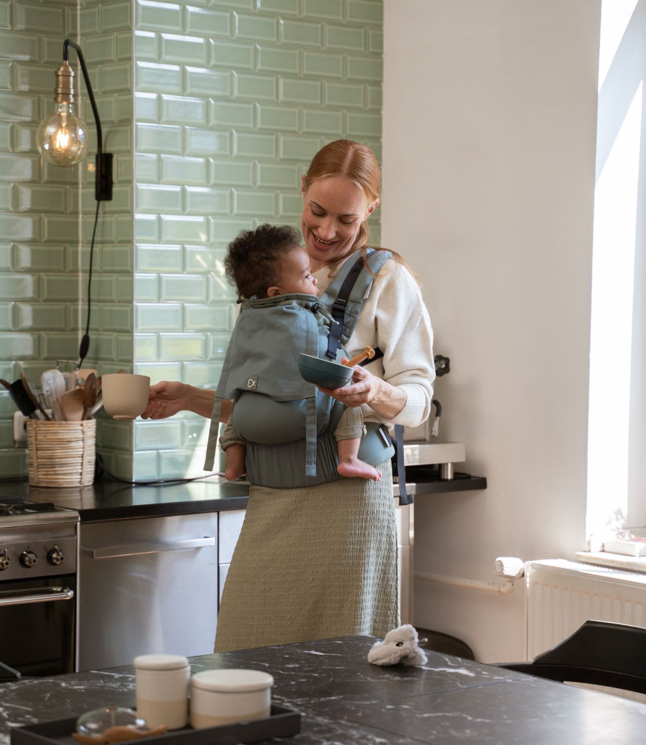 Mutter trägt ihr Baby mit der Fullbuckle in #farbe_krokodilgrün und hat dabei alle Hände frei