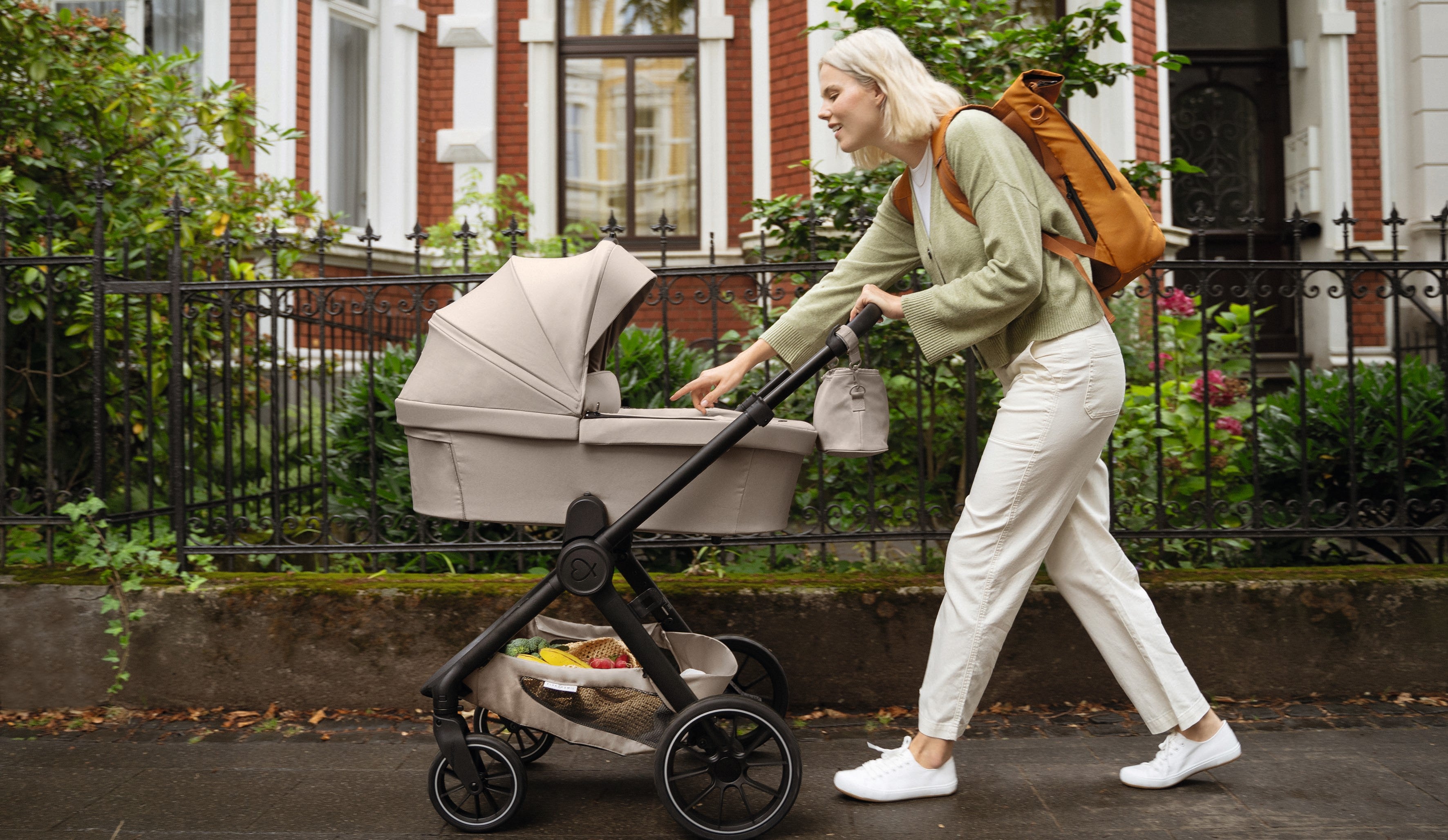 Mutter spaziert mit ihrem Kinderwagen in #farbe_kängurubeige und Rolltop Rucksack in #farbe_fuchsrot