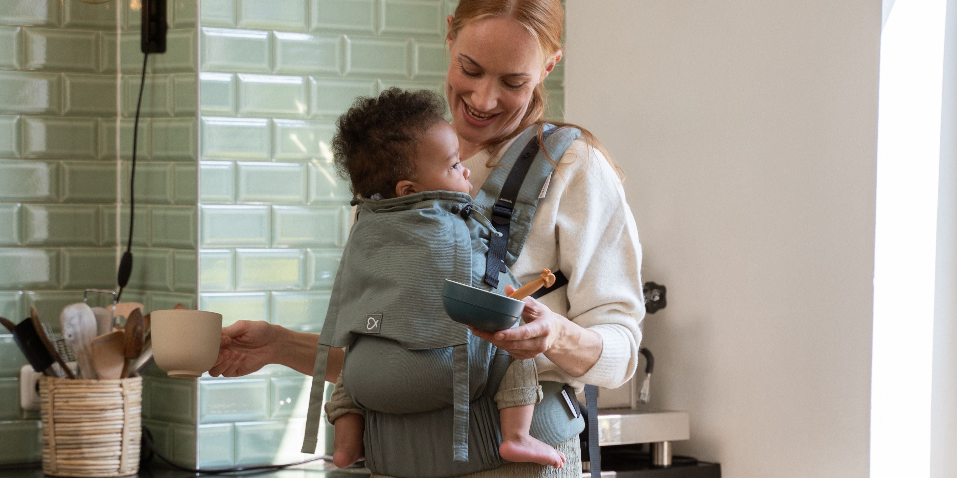 Mutter trägt ihr Baby mit der Fullbuckle in #farbe_krokodilgrün und hat dabei alle Hände frei