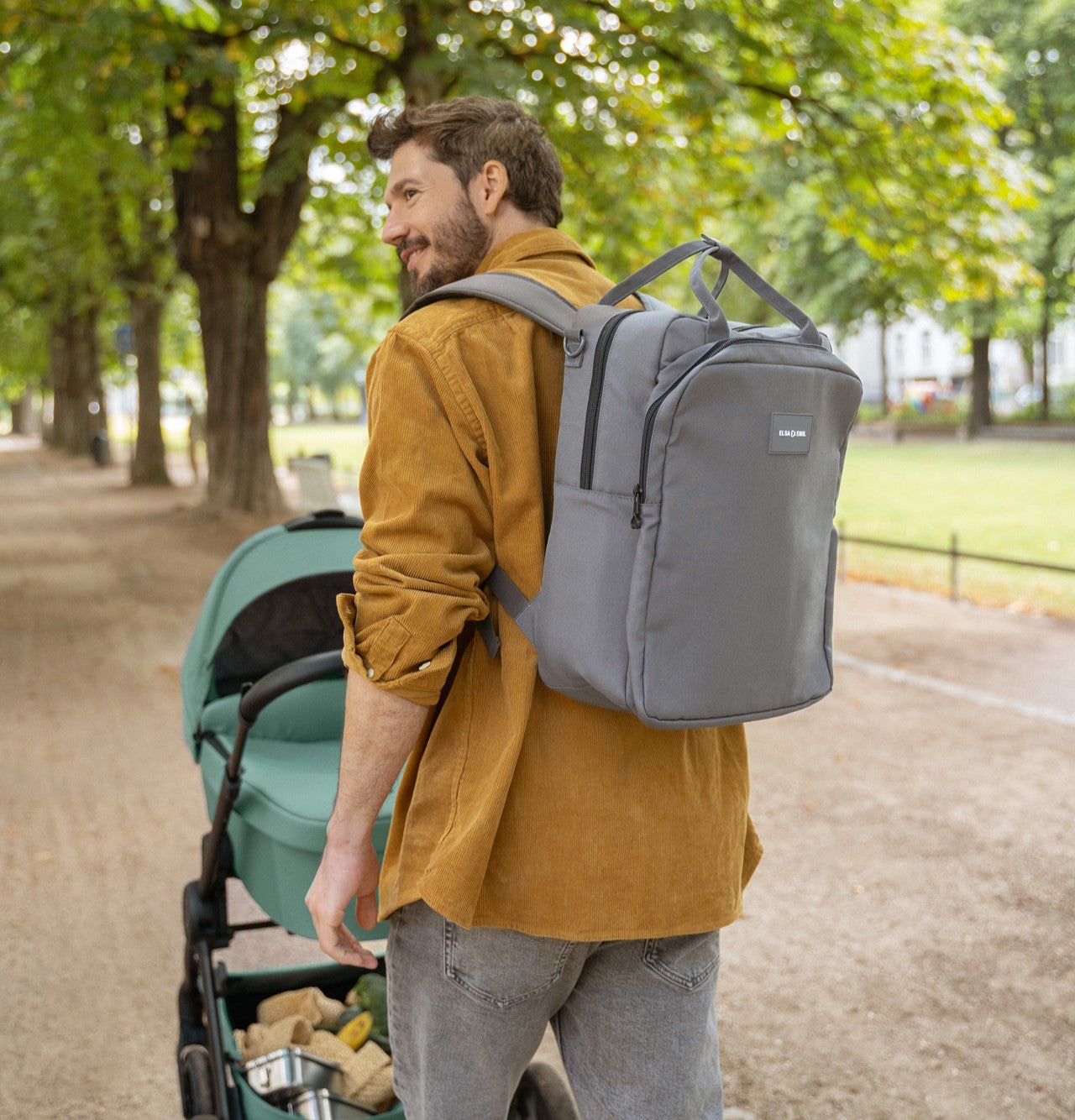 Vater spaziert mit dem Kinderwagen #farbe_krokodilgrün und trägt dabei den Henkel Rucksack #farbe_elefantengrau