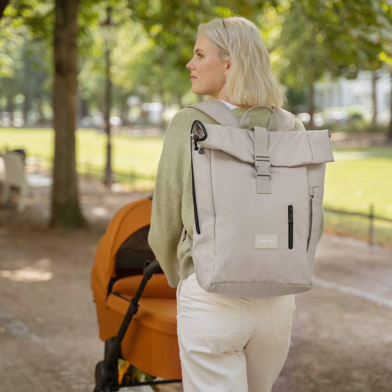 Mutter spaziert mit dem Kindewagen #farbe_fuchsrot, während sie den Rolltop-Rucksack #farbe_kängurubeige trägt